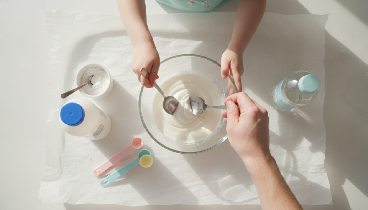 Top-down view of a UK kitchen table set up for slime making: PVA school glue, a mixing bowl, measuring spoons, bicarbonate of soda, saline solution, and a parent helping a child stir the mixture on a parchment-covered table.